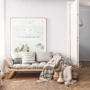 May include: A watercolor print with a light green and blue color scheme featuring a forest scene with the text "You're gonna go far" in black lettering. The print is framed and hanging on a white wall above a light gray couch with a striped throw blanket.