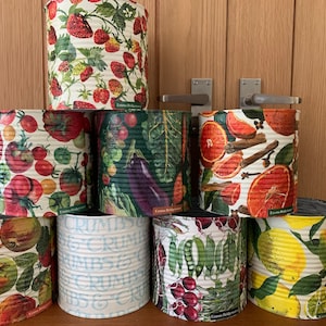 May include: A collection of eight cylindrical metal containers with various colourful designs featuring fruits, vegetables, and other patterns. The containers are stacked and arranged in a row on a wooden surface.