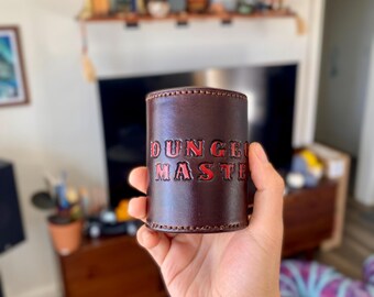May include: A brown leather dice cup with the words "DUNGEON MASTER" embossed in red. The cup is cylindrical and appears to be hand-stitched. The image is taken indoors, with a person holding the cup.