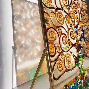 Könnte beinhalten: Ein Buntglaskunstwerk eines Baumes mit wirbelnden Ästen in Gold-, Rot-, Grün- und Blautönen. Das Kunstwerk ist in Gold gerahmt und auf einem goldenen Staffelei ausgestellt.