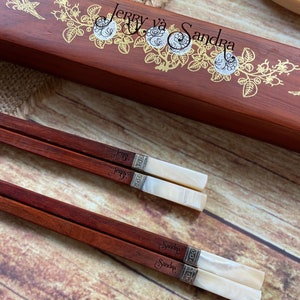May include: Two pairs of dark reddish-brown wooden chopsticks with white pearlescent ends and silver-toned metal bands. One pair is engraved with the name Jerry and the other with Sandra. They are displayed with a matching wooden box featuring gold floral designs and the text Jerry v&agrave; Sandra.