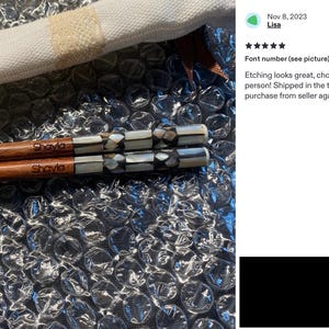 K&ouml;nnte beinhalten: Ein Paar Essst&auml;bchen aus Holz mit dem eingravierten Namen Shayla im braunen Holz und einem dekorativen Griffbereich, der mit einem geometrischen Muster aus schillernden wei&szlig;en und dunkelgrauen Muschelst&uuml;cken eingelegt ist, auf Luftpolsterfolie.