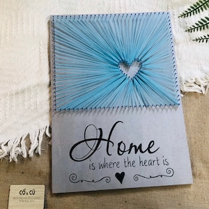 Könnte beinhalten: Ein handgefertigtes String-Art-Stück mit einem hellblauen Herzdesign. Das Kunstwerk enthält den Satz "Home is where the heart is" in schwarzer Schrift. Das Stück ist auf einem grauen Hintergrund montiert.