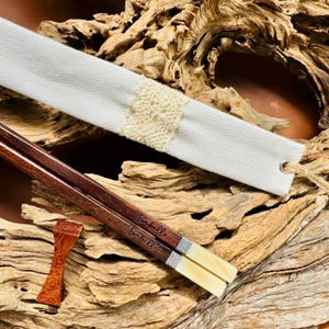 May include: A pair of dark brown wooden chopsticks with the name Isabelle engraved on each, featuring cream-colored ends and silver-toned bands. Included is a white fabric sleeve with a cream lace accent and a small wooden chopstick rest.