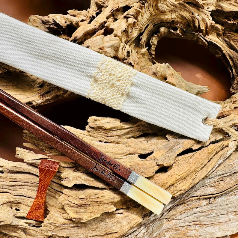 May include: A pair of dark brown wooden chopsticks with the name Isabelle engraved on each, featuring cream-colored ends and silver-toned bands. Included is a white fabric sleeve with a cream lace accent and a small wooden chopstick rest.