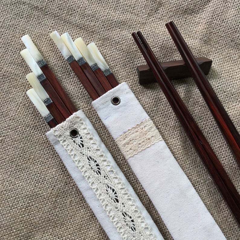 May include: A set of dark brown wooden chopsticks with white shell inlays and silver-toned metal bands, stored in two off-white canvas pouches featuring lace trim and metal grommets, accompanied by a single dark brown wooden chopstick rest.