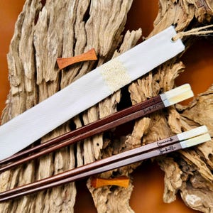 May include: Two pairs of dark brown wooden chopsticks with light-colored tips, each engraved with the names Leonardo and Isabella. The set includes a white fabric sleeve featuring a central cream-colored lace band and a small wooden accent piece.