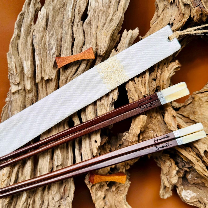May include: Two pairs of dark brown wooden chopsticks with light-colored tips, each engraved with the names Leonardo and Isabella. The set includes a white fabric sleeve featuring a central cream-colored lace band and a small wooden accent piece.