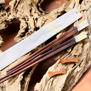 May include: Two pairs of dark brown wooden chopsticks with light-colored tips and engraved names Leonardo and Isabelle. The set includes a white fabric pouch with a textured center detail and two small, reddish-brown wooden chopstick rests.
