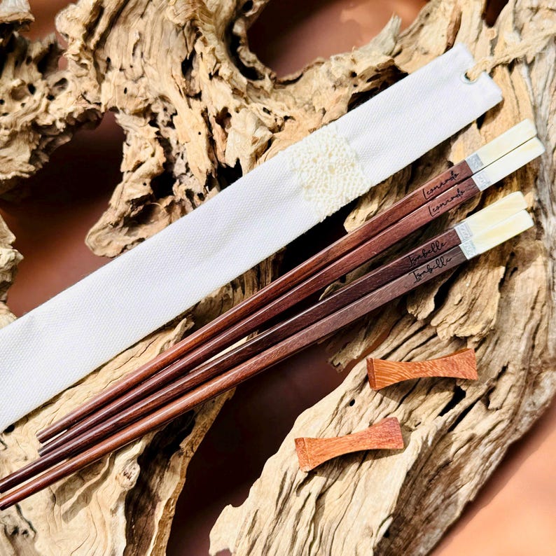 May include: Two pairs of dark brown wooden chopsticks with light-colored tips and engraved names Leonardo and Isabelle. The set includes a white fabric pouch with a textured center detail and two small, reddish-brown wooden chopstick rests.