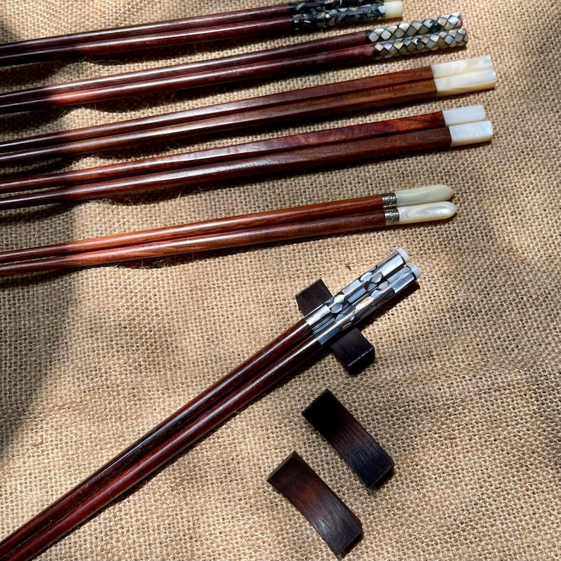 K&ouml;nnte beinhalten: Eine Sammlung von dunkelbraunen Holzst&auml;bchen mit dekorativen Enden, die mit Perlmutt und metallischen Akzenten eingelegt sind. Einige Paare haben wei&szlig;e Muschelspitzen, andere gemusterte Metalleinlagen. Zwei dunkle Holzablagen sind ebenfalls enthalten.