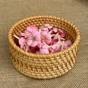 Può includere: Un cesto intrecciato in vimini pieno di fiori di stoffa rosa. Il cesto è rotondo e di colore marrone naturale.