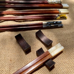 May include: A collection of dark brown wooden chopsticks featuring decorative ends with inlaid white, black, and metallic patterns. Two dark brown wooden chopstick rests are positioned nearby, with one pair of chopsticks resting on a third wooden rest.
