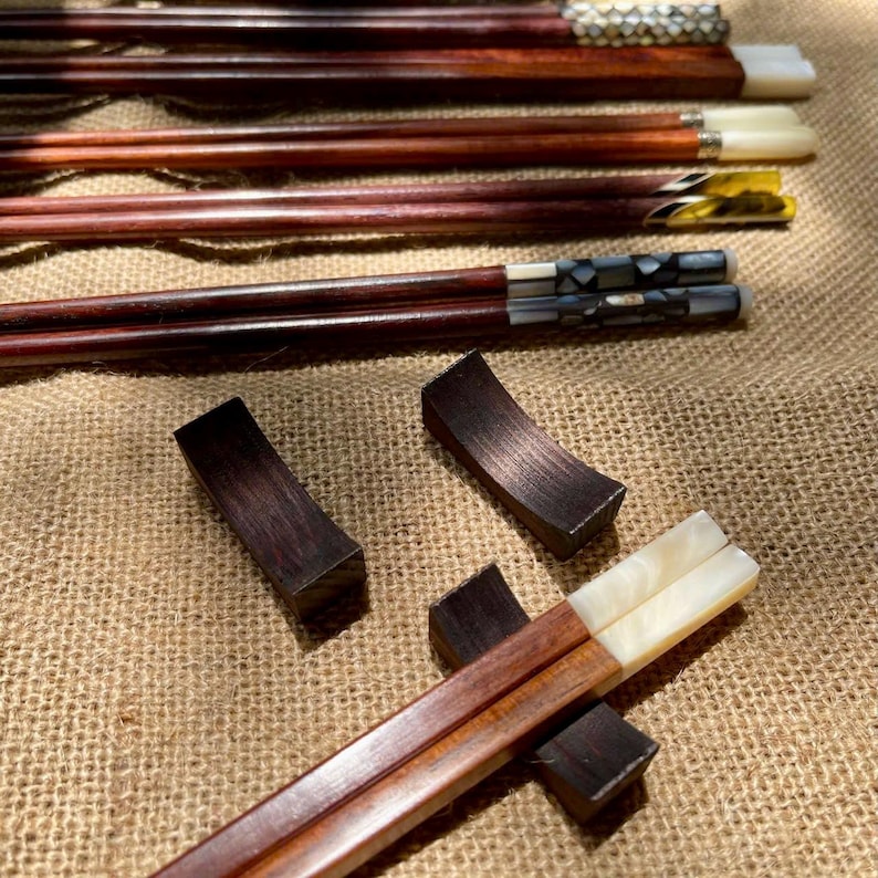 May include: A collection of dark brown wooden chopsticks featuring decorative ends with inlaid white, black, and metallic patterns. Two dark brown wooden chopstick rests are positioned nearby, with one pair of chopsticks resting on a third wooden rest.