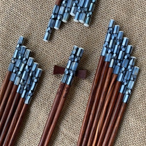 K&ouml;nnte beinhalten: Eine Sammlung von Holzst&auml;bchen mit dunkelbraunen Sch&auml;ften und Griffen, die mit schillernden grauen und wei&szlig;en Perlmutt-Mosaikfliesen eingelegt sind. Ein Paar ruht auf einer dunkelbraunen Holzablage. Der Text "Fancy Gift" ist in das Holz eingraviert.