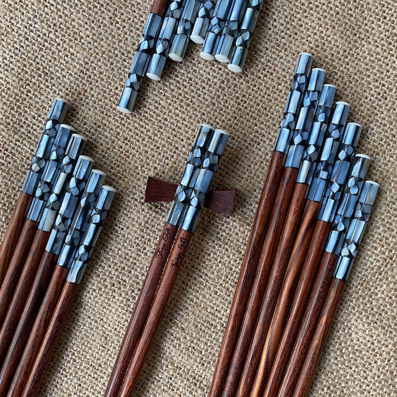 K&ouml;nnte beinhalten: Eine Sammlung von Holzst&auml;bchen mit dunkelbraunen Sch&auml;ften und Griffen, die mit schillernden grauen und wei&szlig;en Perlmutt-Mosaikfliesen eingelegt sind. Ein Paar ruht auf einer dunkelbraunen Holzablage. Der Text "Fancy Gift" ist in das Holz eingraviert.