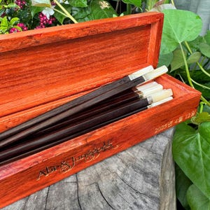 May include: A set of dark brown chopsticks with light-colored tips resting inside a rectangular orange-red wooden box. The box features gold engraved text that reads "Abr & Jungkook" and the date "2025년 4월 12일" on the front edge.