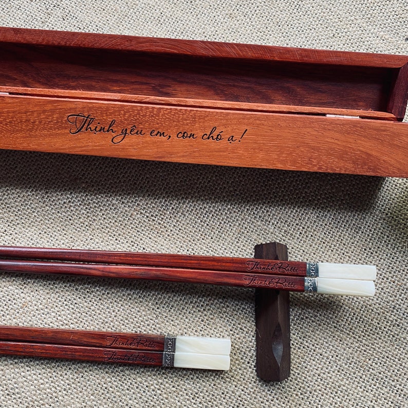 May include: A pair of dark brown wooden chopsticks with white tips and silver-toned metal accents, engraved with the text "Thinh&Ritti". The chopsticks rest on a dark wooden holder next to a matching wooden box engraved with "Thịnh y&ecirc;u em, con ch&oacute; ạ!".