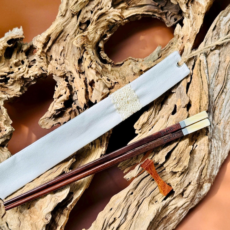 May include: A pair of dark brown wooden chopsticks with light-colored tips, featuring the name Isabella engraved on the handles. The set includes a white fabric sleeve with a central cream-colored lace detail and a small, orange-brown wooden rest.