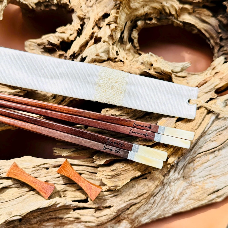 May include: Two pairs of dark brown wooden chopsticks with light-colored tips and silver-toned metal accents. Each pair is engraved with the names Leonardo and Isabelle. A white fabric sleeve with a lace detail and two small wooden rests.