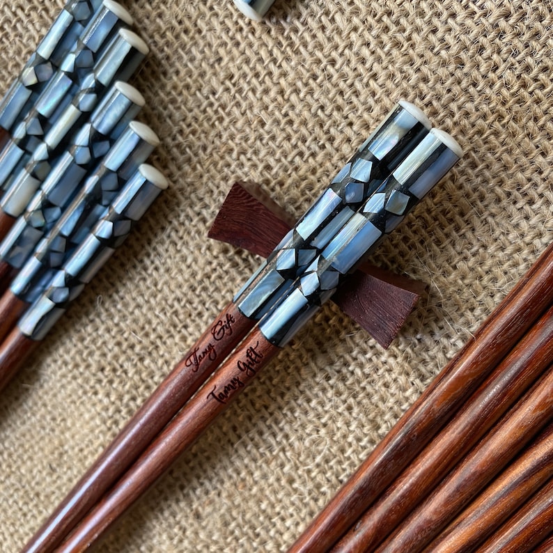 K&ouml;nnte beinhalten: Ein Set aus braunen Holzst&auml;bchen mit einer dekorativen Einlage aus irisierenden, geometrischen Perlmuttst&uuml;cken am oberen Ende. Der Schriftzug Tamy Gift ist in den Griff jedes St&auml;bchens eingraviert. Das Set enth&auml;lt eine braune Holzablage.