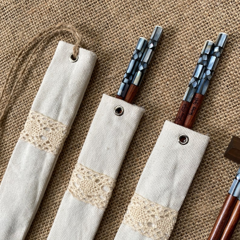 K&ouml;nnte beinhalten: Drei Paar Essst&auml;bchen aus Holz mit dunkelbraunen Sch&auml;ften und Griffen, die mit schillernden grauen geometrischen Perlmuttpl&auml;ttchen eingelegt sind. Jedes Paar steckt in einer cremefarbenen Stoffh&uuml;lle mit Metall&ouml;se und Spitzenbesatz.