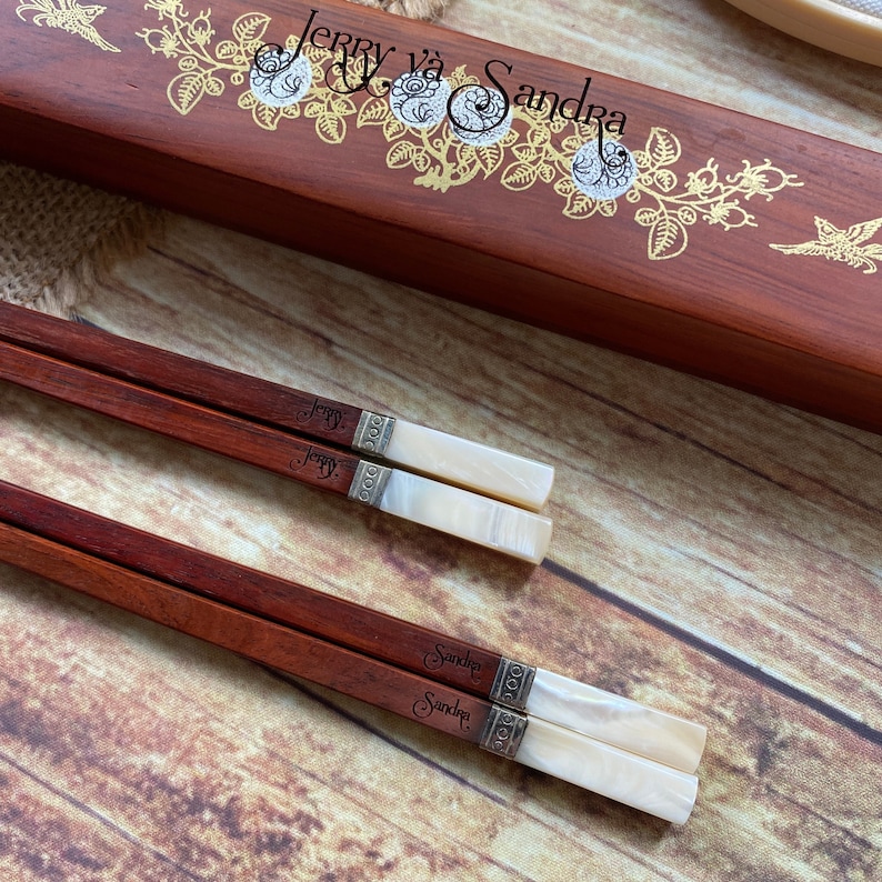 May include: Two pairs of dark reddish-brown wooden chopsticks with white pearlescent ends and silver-toned metal bands. One pair is engraved with the name Jerry and the other with Sandra. They are displayed with a matching wooden box featuring gold floral designs and the text Jerry v&agrave; Sandra.