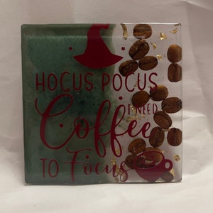 May include: A square resin coaster featuring a green marbled section and a clear section embedded with whole roasted coffee beans and gold flakes. The text HOCUS POCUS I NEED COFFEE TO FOCUS is printed in red along with a witch hat and coffee cup icon.
