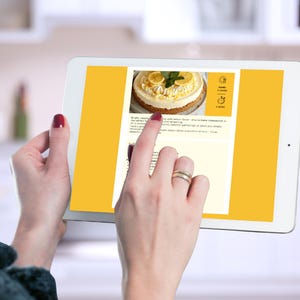 Hands viewing the Lemon Cheesecake recipe page on a tablet in a kitchen setting.