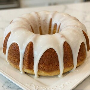 May include: A golden-brown bundt cake with white icing drizzled over the top. The cake sits on a white, square, decorative plate. The icing drips down the sides of the cake, creating a visually appealing dessert.