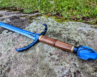 Puede incluir: Una espada de fantasía con una hoja azul y un mango de madera. La guarda presenta detalles curvos en azul oscuro y un anillo dorado. El pomo es azul con un diseño texturizado. La espada descansa sobre una roca musgosa.