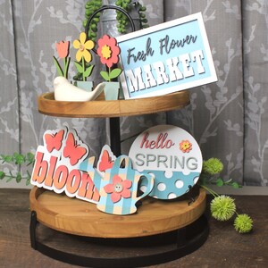 May include: Two-tiered wooden stand with a black metal base. The top tier features a blue and white sign that says "Fresh Flower Market". The bottom tier has a variety of spring-themed decorations, including a watering can, butterflies, and a sign that says "hello spring".