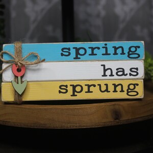 May include: A wooden sign with three stacked blocks painted in blue, white, and yellow. The sign reads "spring has sprung" with a small wooden flower decoration.