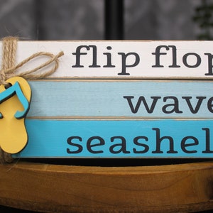 May include: A wooden sign with three stacked blocks. The top block is white with the text "flip flops" in black. The middle block is light blue with the text "waves" in black. The bottom block is blue with the text "seashells" in black. A yellow wooden flip flop is attached to the sign with twine.