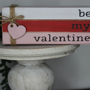 May include: A stack of three wooden blocks painted in white, red, and pink with the words "be my valentine" painted in black. A pink heart is attached to the blocks with twine.