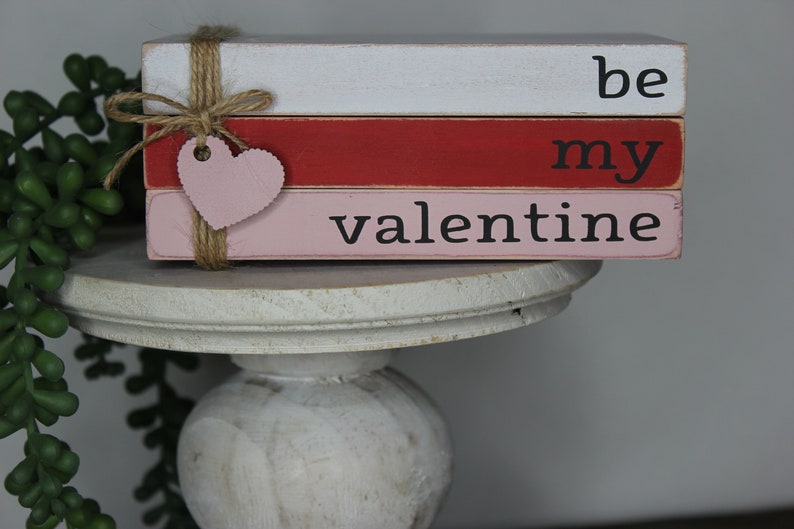 May include: A stack of three wooden blocks painted in white, red, and pink with the words "be my valentine" painted in black. A pink heart is attached to the blocks with twine.