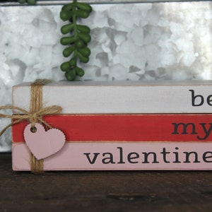 May include: Stacked wooden blocks painted in pink, red, and white with the words "be my valentine" written in black. The blocks are tied together with twine and a pink heart-shaped tag.