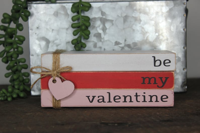 May include: Stacked wooden blocks painted in pink, red, and white with the words "be my valentine" written in black. The blocks are tied together with twine and a pink heart-shaped tag.