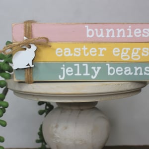 May include: A stack of three wooden blocks painted in pink, yellow, and green with the words "bunnies", "easter eggs", and "jelly beans" painted in white. A white wooden bunny cutout is attached to the blocks with twine.