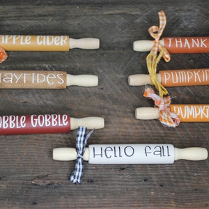 May include: Five wooden rolling pins decorated with fall-themed phrases and tied with ribbons. The phrases are "Apple Cider", "Hayrides", "Gobble Gobble", "Pumpkin Pie", "Pumpkin Spice", and "Hello Fall".
