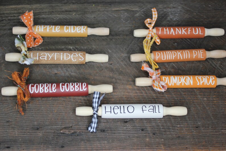 May include: Five wooden rolling pins decorated with fall-themed phrases and tied with ribbons. The phrases are "Apple Cider", "Hayrides", "Gobble Gobble", "Pumpkin Pie", "Pumpkin Spice", and "Hello Fall".