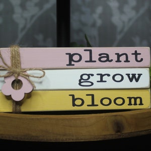 May include: Three wooden blocks stacked on top of each other. The top block is pink and says "plant", the middle block is white and says "grow", and the bottom block is yellow and says "bloom". A pink flower cutout is tied to the blocks with twine.