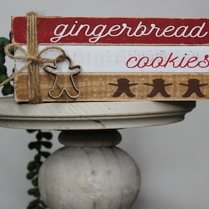 May include: A wooden sign with the words "gingerbread cookies" painted on it. The sign is decorated with gingerbread man cutouts and a brown twine bow.