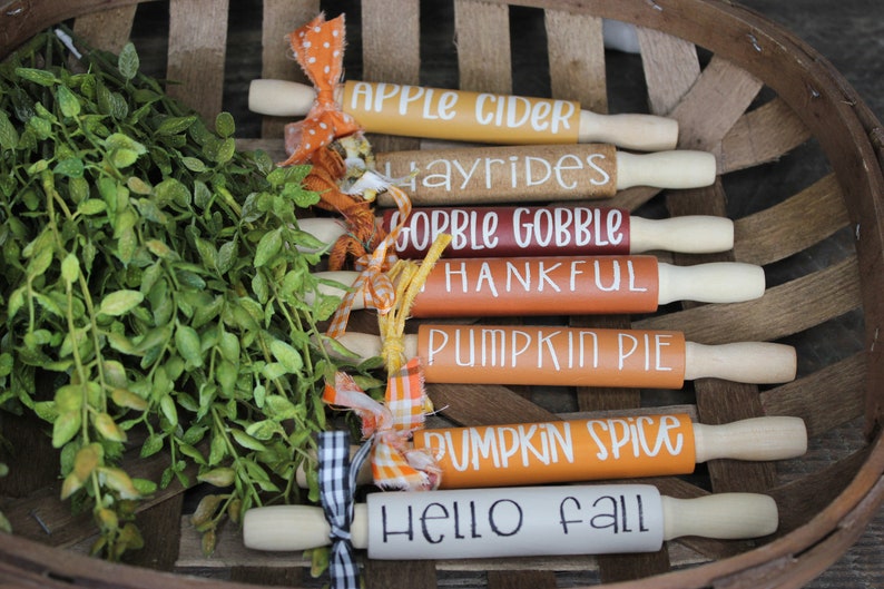 May include: A set of seven wooden rolling pins decorated with fall-themed phrases, including "Apple Cider", "Hayrides", "Gobble Gobble", "Thankful", "Pumpkin Pie", "Pumpkin Spice", and "Hello Fall". The rolling pins are arranged in a basket with green foliage.