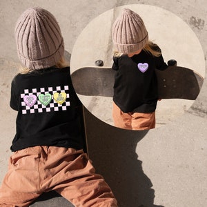 Może przedstawiać: Dziecko w czarnym t-shircie w kratkę z kolorowymi serduszkami z cukierk&oacute;w, na kt&oacute;rych napisane jest "Pretty" i "Sweet". Dziecko ma na sobie brązowe spodnie i białe buty sportowe i trzyma deskorolkę.