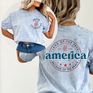 May include: A light blue t-shirt with a distressed, marbled effect. The front of the shirt has a small graphic with the text "Grove 2023 Memories". The back of the shirt has a large graphic with the text "Land of the Free, Since 1776, America, Because of the Brave".