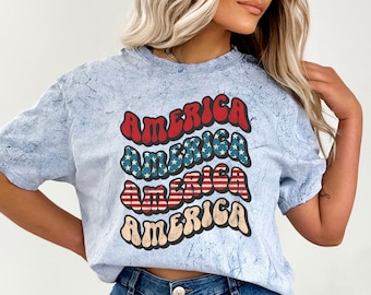 May include: A light blue t-shirt with a tie-dye effect featuring the word "America" repeated four times in a retro font with red, white, and blue stars and stripes.