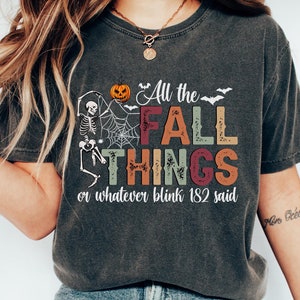 May include: Dark gray t-shirt with the text "All the FALL THINGS or whatever blink 182 said." The shirt features a skeleton, pumpkin, and spiderweb graphic, perfect for the fall season. The shirt is paired with blue jeans and white sneakers.