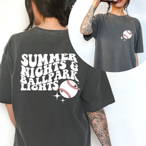 Pu&ograve; includere: Una maglietta oversize di colore grigio scuro con una grafica bianca che dice "Summer Nights & Ballpark Lights" con un grafico di una palla da baseball.