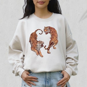 May include: A cream colored sweatshirt with a graphic of two tigers facing each other. The tigers are orange and black with white stripes.